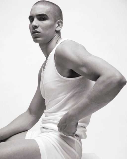 A man with a shaved head sits against a white background in underwear and a tank top, showcasing a minimalist fashion aesthetic.