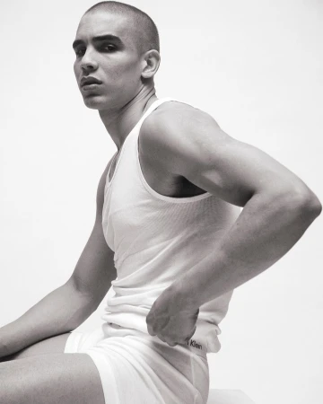 A man with a shaved head sits against a white background in underwear and a tank top, showcasing a minimalist fashion aesthetic.