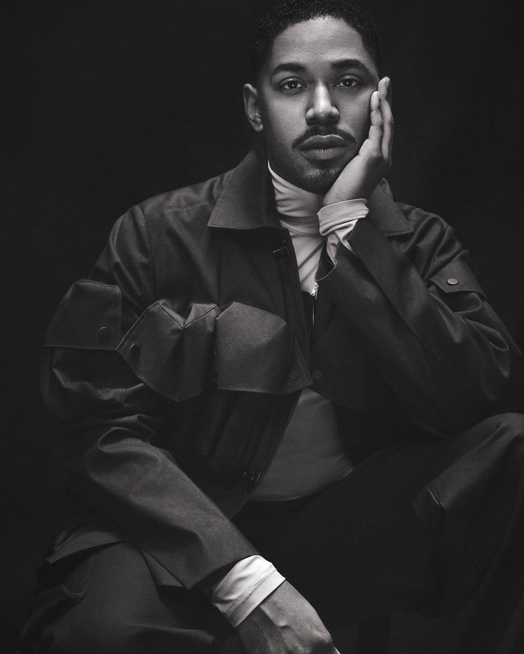 Portrait of a Black man with a mustache wearing a turtleneck and utility jacket, photographed by Isaac Anthony in monochrome.