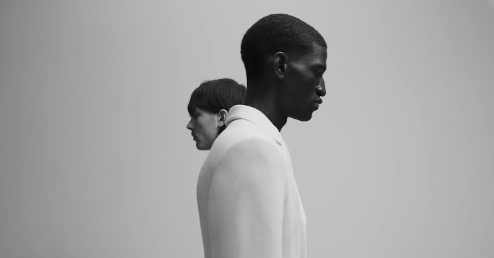 Monochrome portrait of a Black man and a White woman, heads close together, both wearing white jackets against a light grey backdrop.