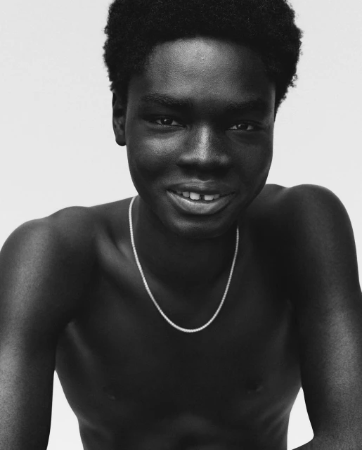 Monochrome portrait of a Black adult male with natural hair, smiling, wearing a silver necklace against a white background.