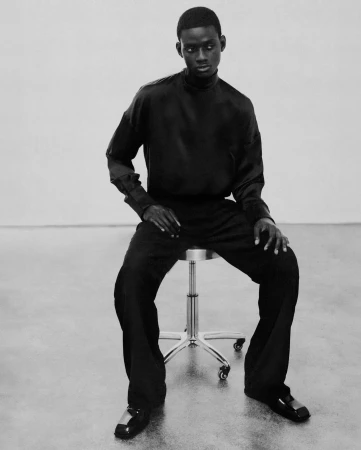 Black and white portrait of a black man wearing dark attire while seated on a metal stool against a plain studio background.