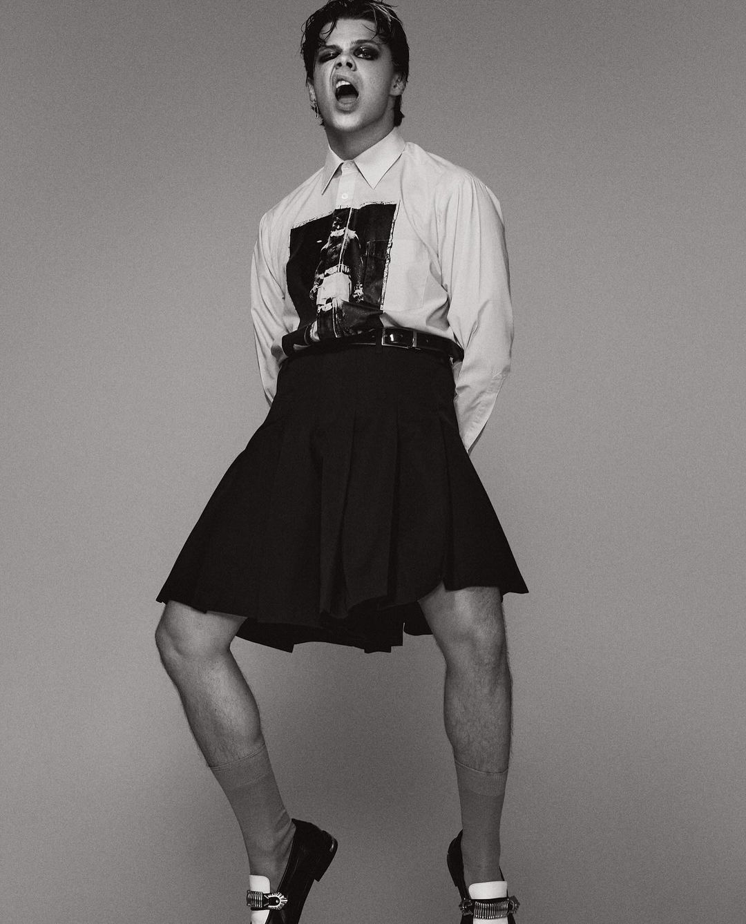 Monochrome portrait of a slender male wearing a black skirt, shirt, and loafers, mouth open and jumping indoors in a photo studio.