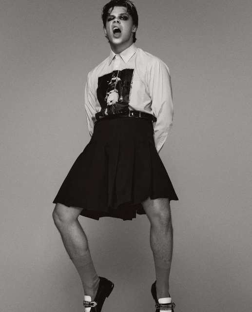 Monochrome portrait of a slender male wearing a black skirt, shirt, and loafers, mouth open and jumping indoors in a photo studio.