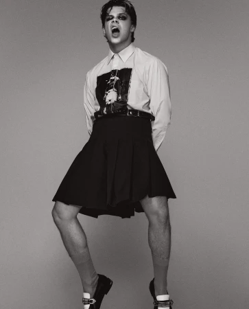 Monochrome portrait of a slender male wearing a black skirt, shirt, and loafers, mouth open and jumping indoors in a photo studio.