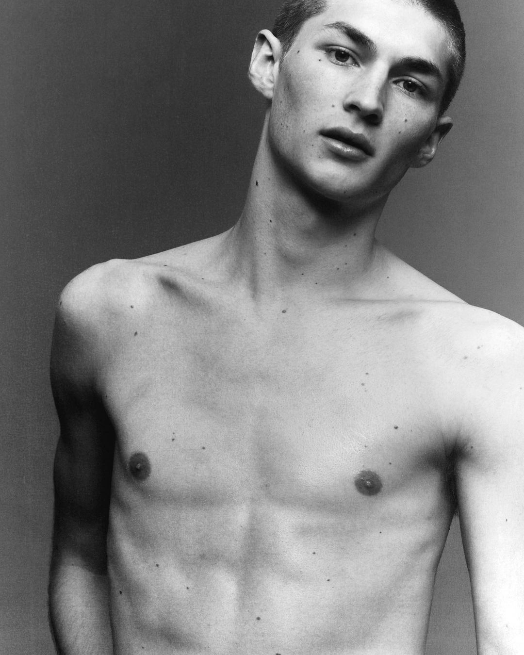 Portrait of a shirtless young man with short hair and freckles against a grey background by Isaac Anthony