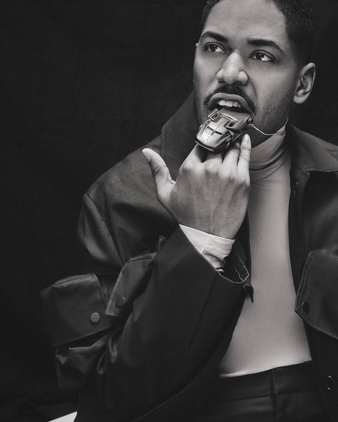 Monochrome portrait of a black man wearing a turtleneck and jacket, biting a toy car held by his finger.