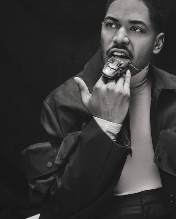 Monochrome portrait of a black man wearing a turtleneck and jacket, biting a toy car held by his finger.