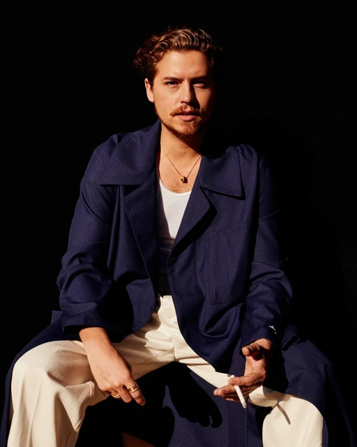 A portrait of a blonde man wearing a navy coat and cream-colored pants. He is sitting indoors against a dark background.