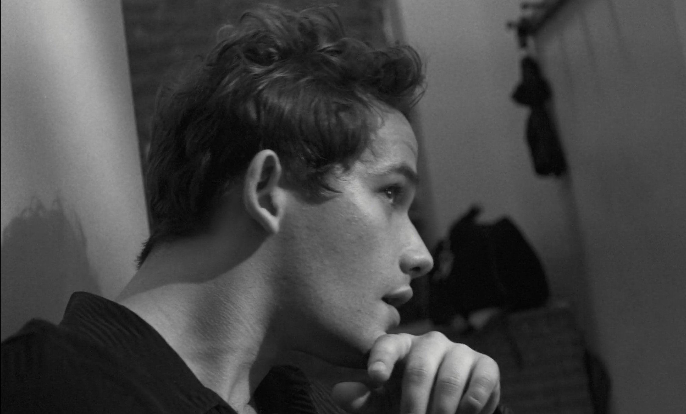Black and white portrait of a young adult male with dark curly hair, hand to chin, looking to the side in an indoor setting.