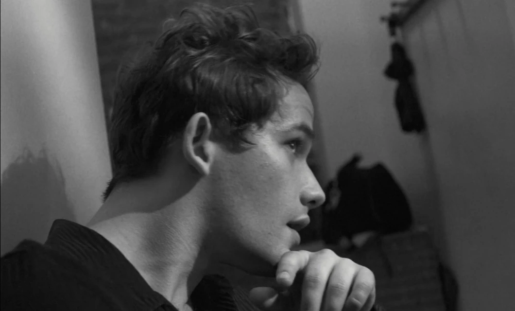 Black and white portrait of a young adult male with dark curly hair, hand to chin, looking to the side in an indoor setting.