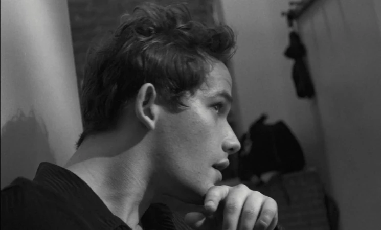 Black and white portrait of a young adult male with dark curly hair, hand to chin, looking to the side in an indoor setting.