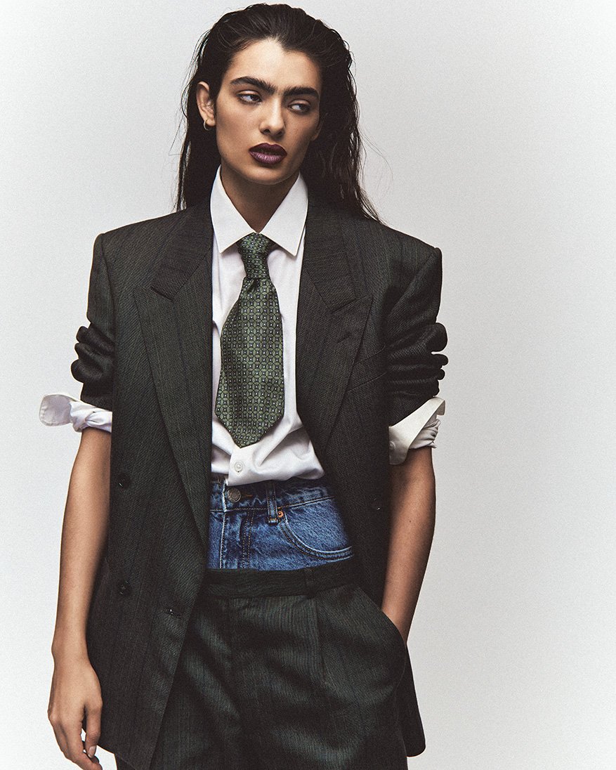 A woman in a business suit and denim shorts poses in a studio with a white background, showcasing a blend of formal and casual fashion.