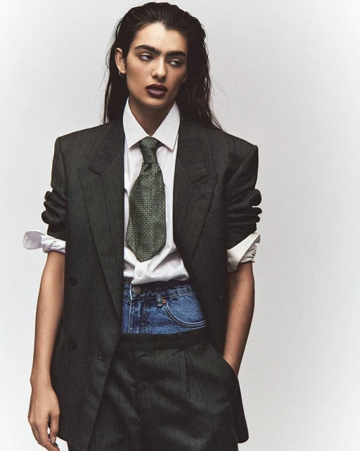 A woman in a business suit and denim shorts poses in a studio with a white background, showcasing a blend of formal and casual fashion.