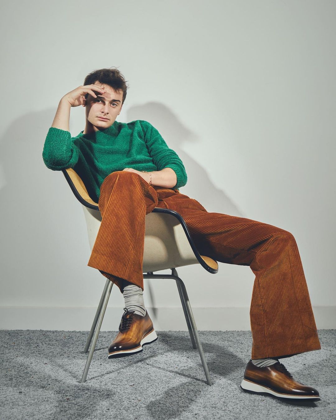 Young man poses in green sweater and orange corduroy pants, seated on mid-century modern chair, wearing leather dress shoes on grey carpet.