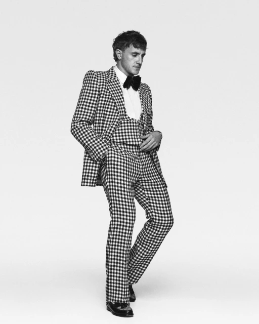 Portrait of a man in a checkered suit, posing in a black and white studio setting. He is wearing a bow tie and dress shoes.