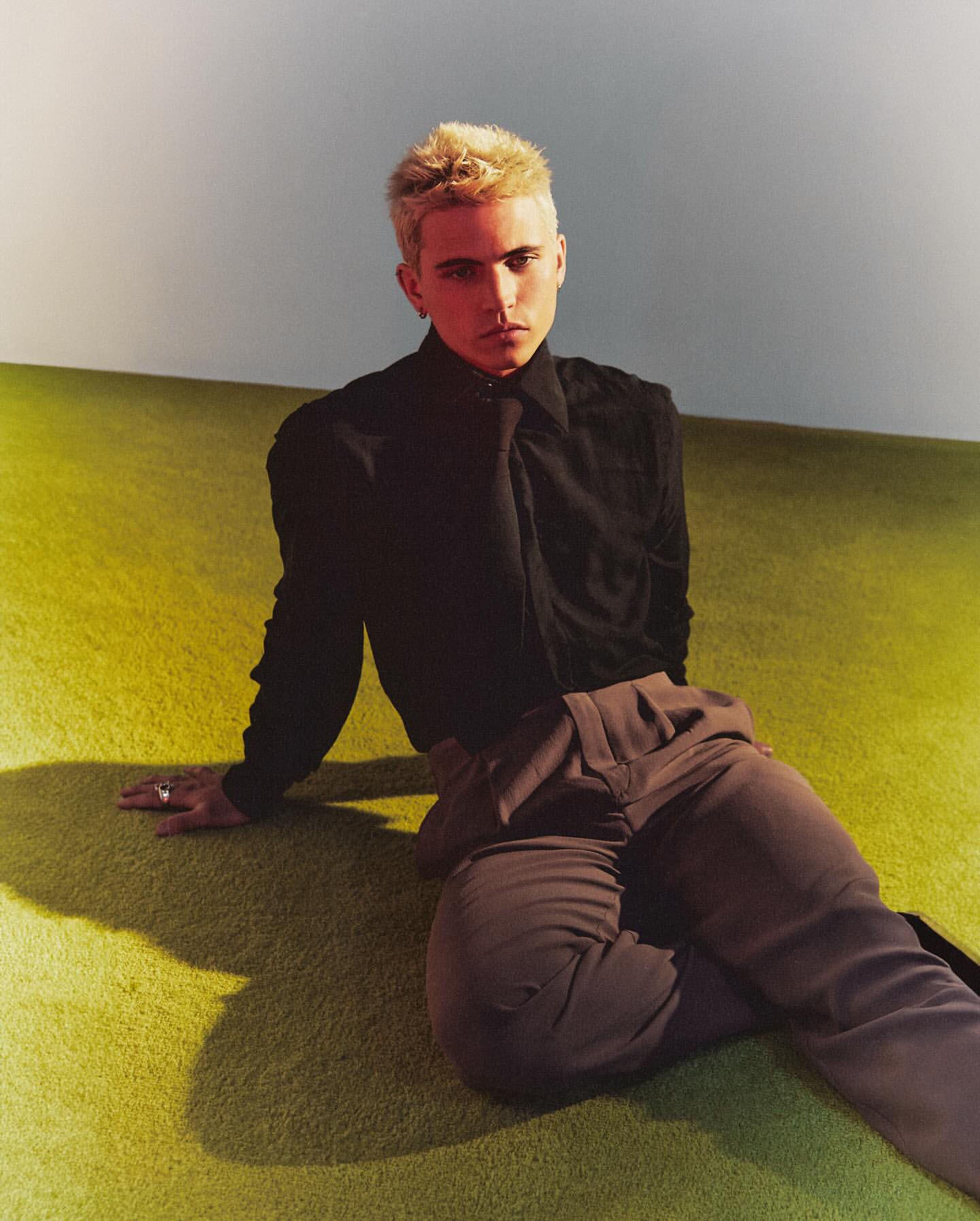 Portrait of a young adult with blonde hair sitting on a green carpet in front of a white wall, wearing a black shirt and brown pants.