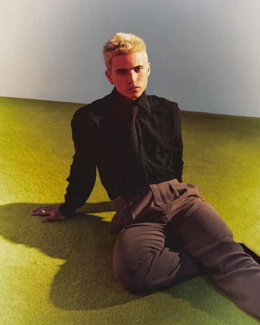Portrait of a young adult with blonde hair sitting on a green carpet in front of a white wall, wearing a black shirt and brown pants.
