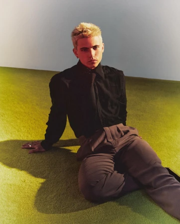 Portrait of a young adult with blonde hair sitting on a green carpet in front of a white wall, wearing a black shirt and brown pants.