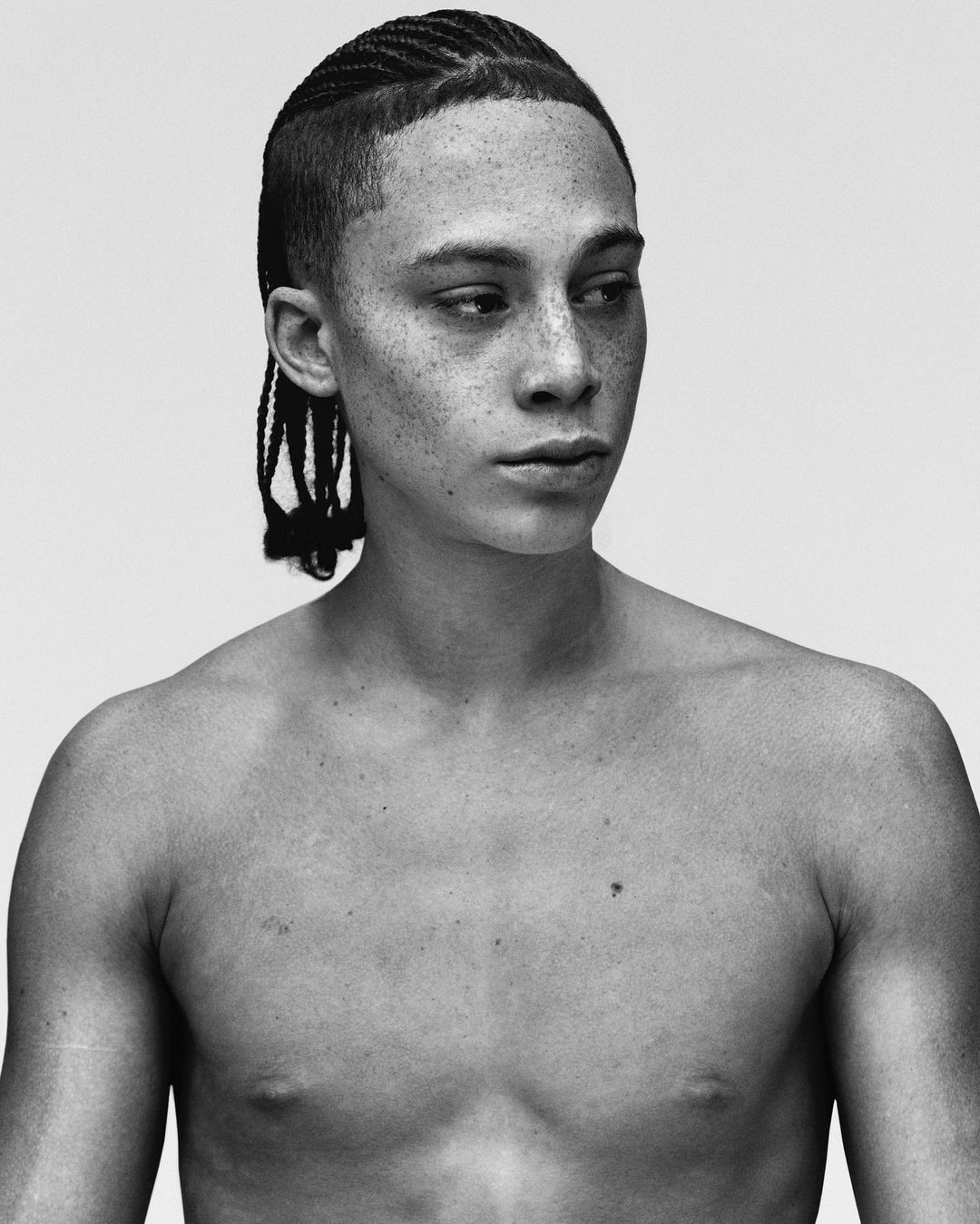 Striking black and white portrait of a topless young man with freckles and cornrow braids, captured in a minimalist studio setting.