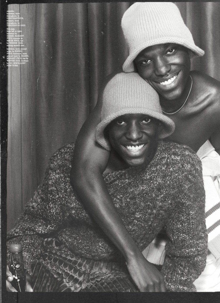 Two smiling Black men wearing knitted bucket hats. One wears a sweater and sits while the other stands behind him with his arm around him.