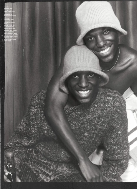 Two smiling Black men wearing knitted bucket hats. One wears a sweater and sits while the other stands behind him with his arm around him.