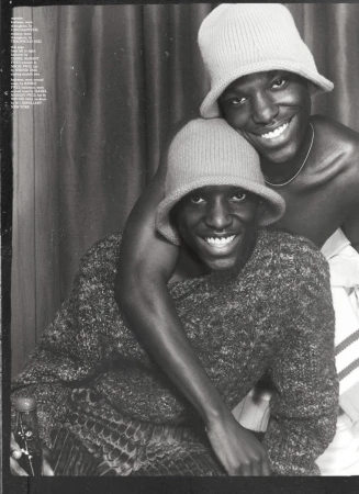 Two smiling Black men wearing knitted bucket hats. One wears a sweater and sits while the other stands behind him with his arm around him.