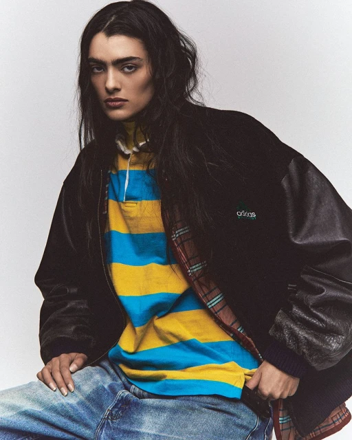 Portrait of a fashion model wearing a black varsity jacket over a striped polo shirt and jeans, posing in a photo studio.