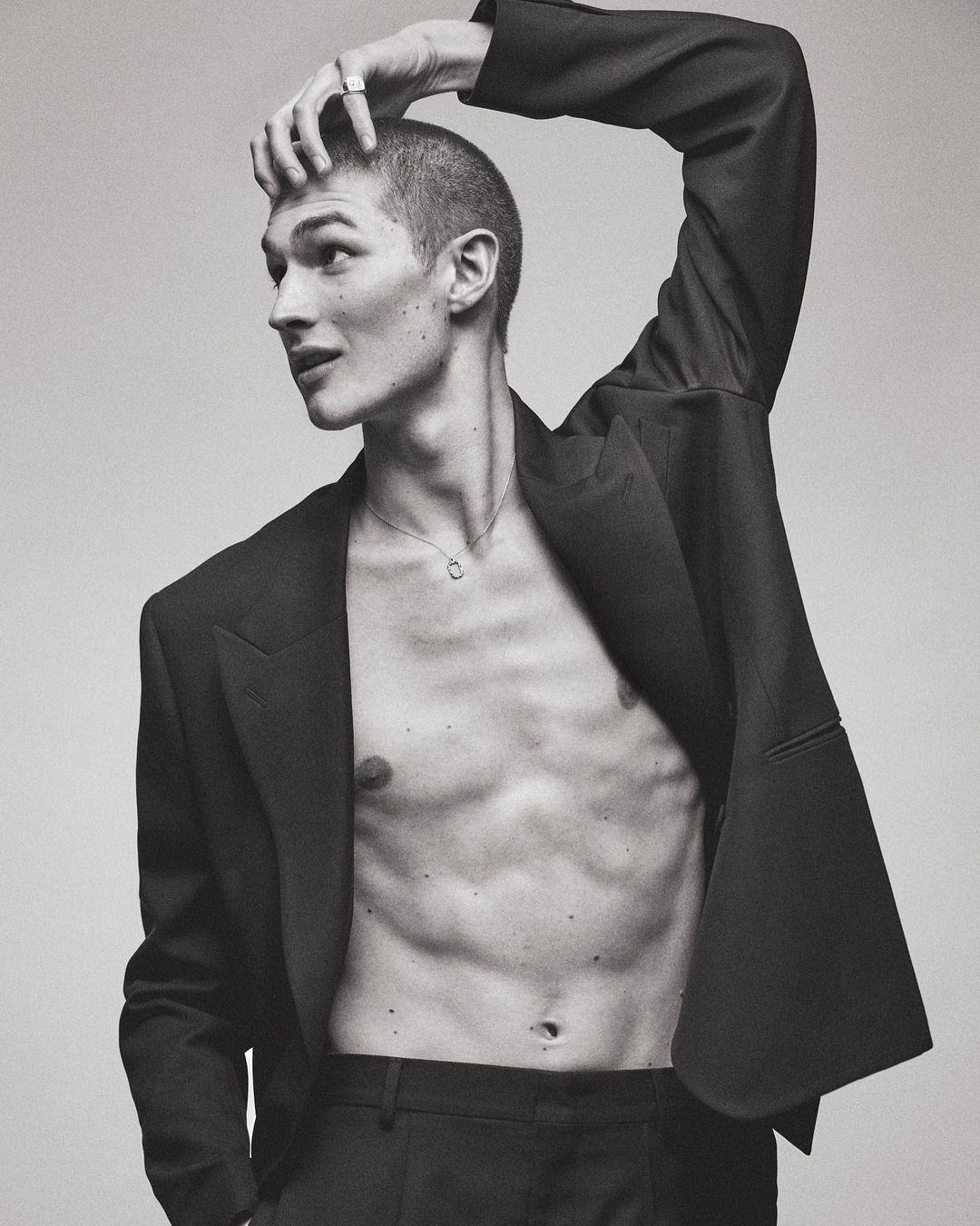 Monochromatic portrait of a shirtless male model in a suit jacket posing with his hand on his head in a bright studio setting.