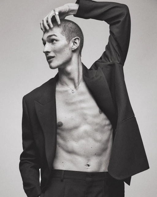 Monochromatic portrait of a shirtless male model in a suit jacket posing with his hand on his head in a bright studio setting.