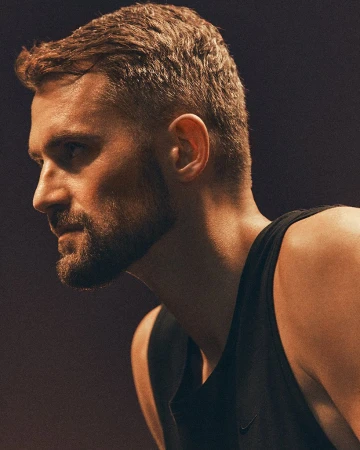 Profile portrait of a bearded man in his 30s wearing a black tank top under warm directional lighting.
