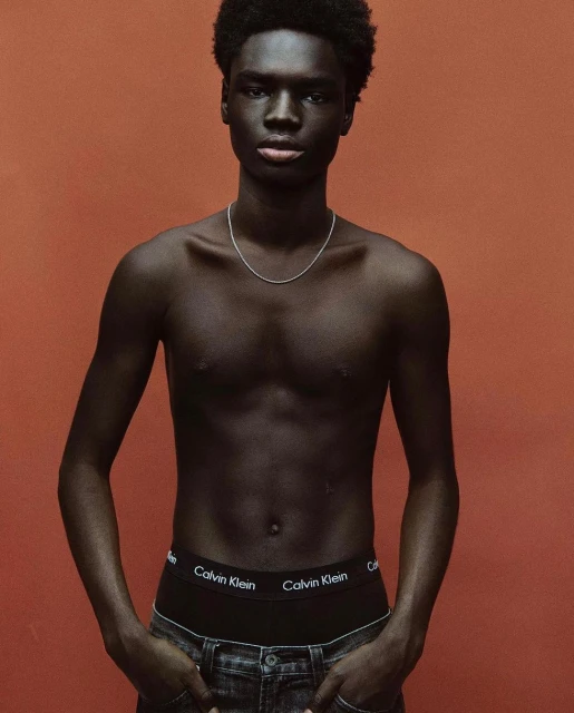 Portrait of a young Black man with bare torso, wearing Calvin Klein underwear and dark jeans, against a solid orange backdrop.