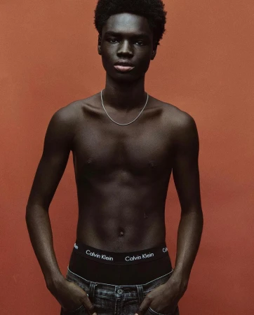 Portrait of a young Black man with bare torso, wearing Calvin Klein underwear and dark jeans, against a solid orange backdrop.