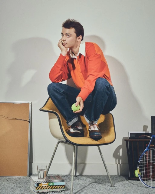 Young man in preppy fashion poses in a photo studio while sitting on a mid-century modern chair and holding a tennis ball.