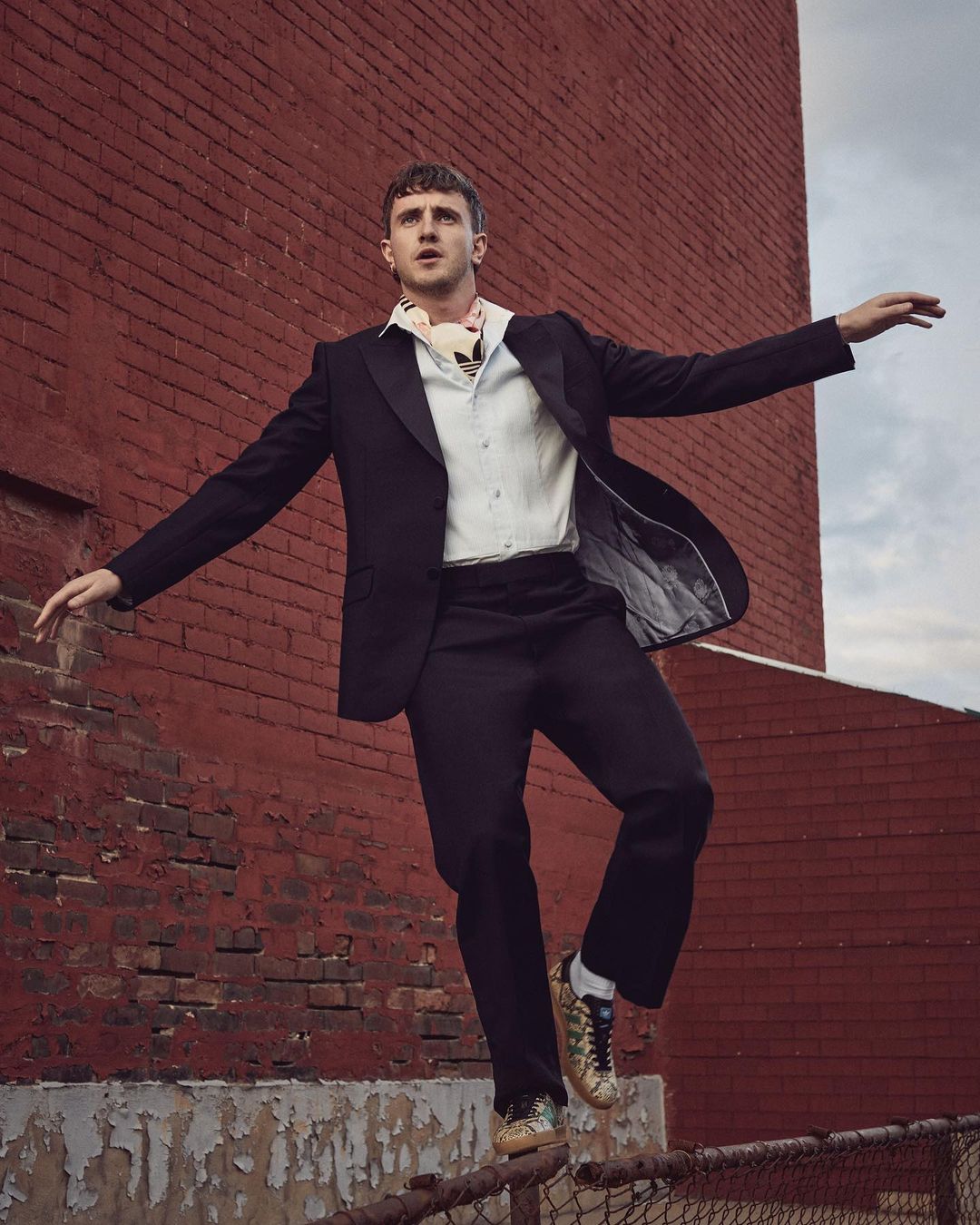 An adult male fashion model balances on a chain link fence wearing a black suit with a white shirt in front of a brick wall.