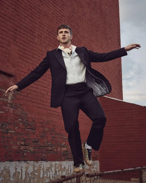 An adult male fashion model balances on a chain link fence wearing a black suit with a white shirt in front of a brick wall.