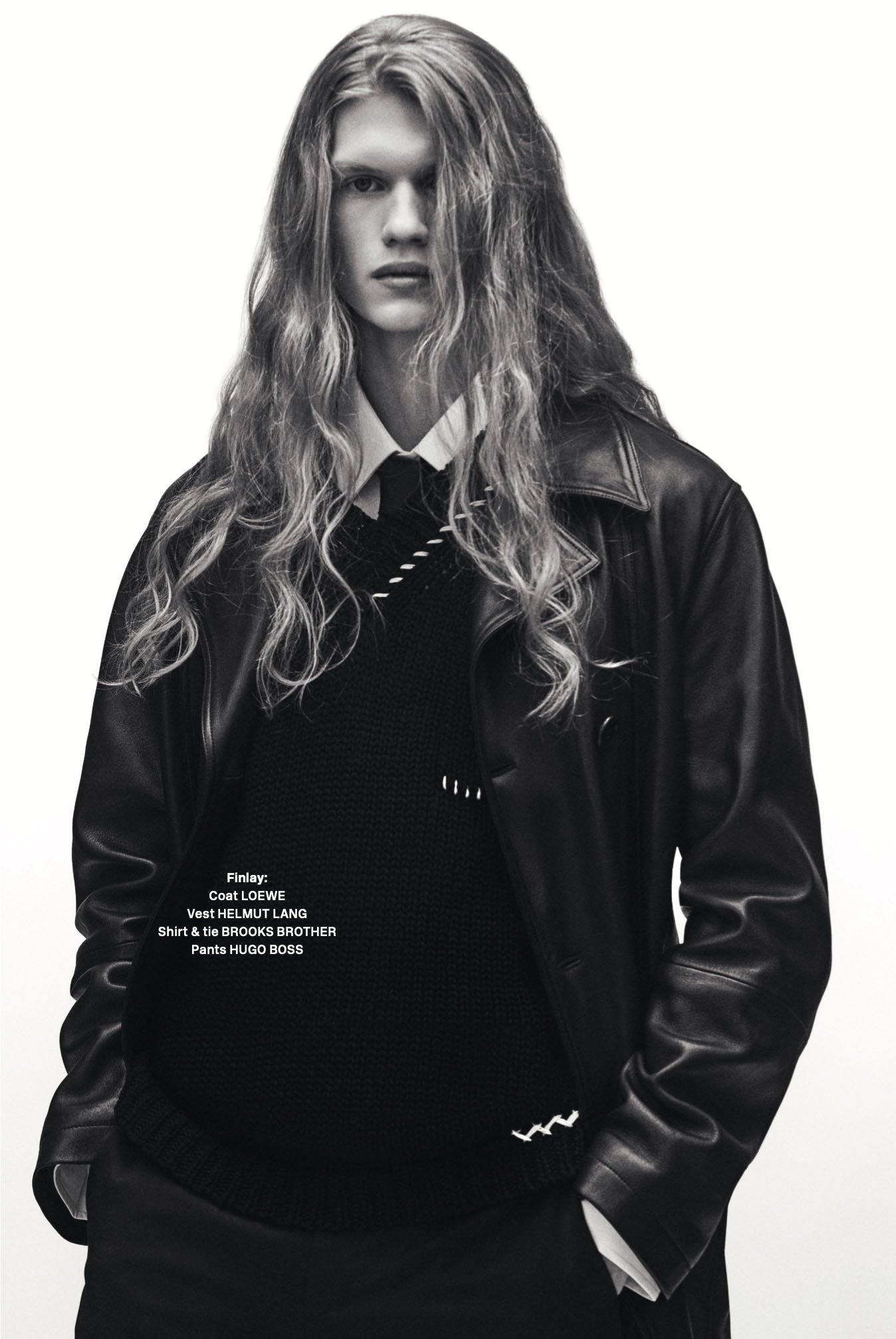 Black and white portrait of a male model with long wavy hair wearing a leather coat, vest, and tie against a plain white background.