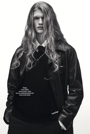 Black and white portrait of a male model with long wavy hair wearing a leather coat, vest, and tie against a plain white background.