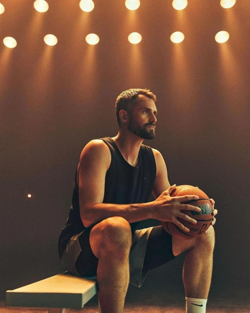 Bearded man in a black tank top sits indoors holding a basketball, with the backlights creating a dramatic ambiance.