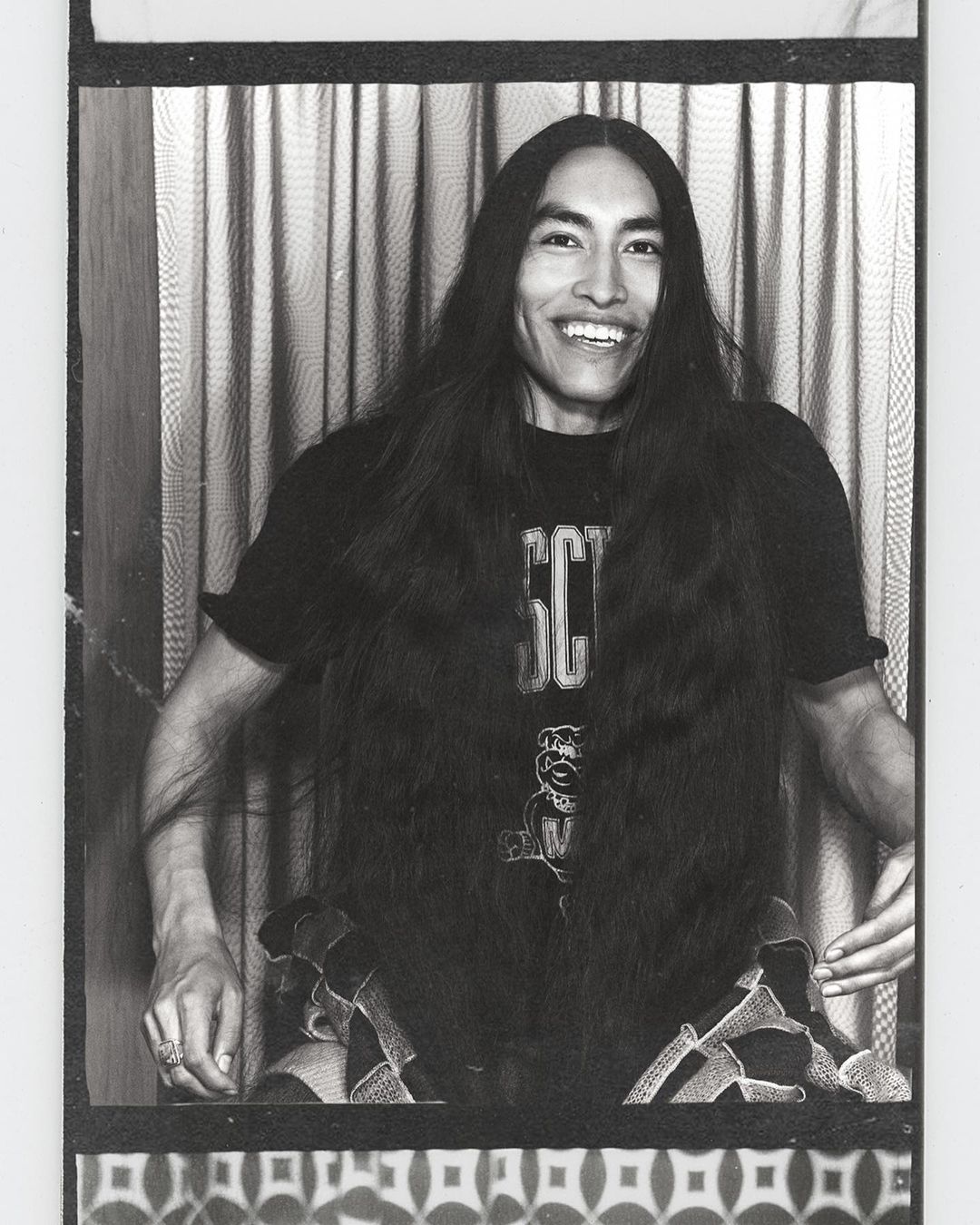 Portrait of a smiling adult male with long dark hair wearing a graphic tee shirt, seated in front of a patterned background.