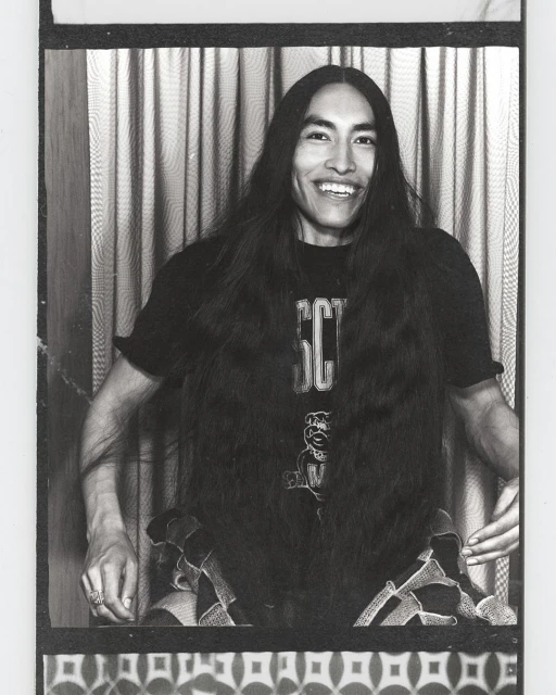 Portrait of a smiling adult male with long dark hair wearing a graphic tee shirt, seated in front of a patterned background.