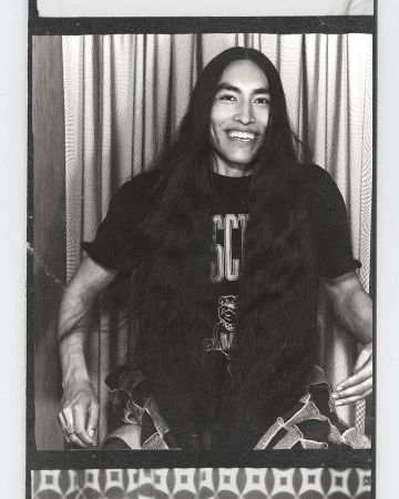 Portrait of a smiling adult male with long dark hair wearing a graphic tee shirt, seated in front of a patterned background.