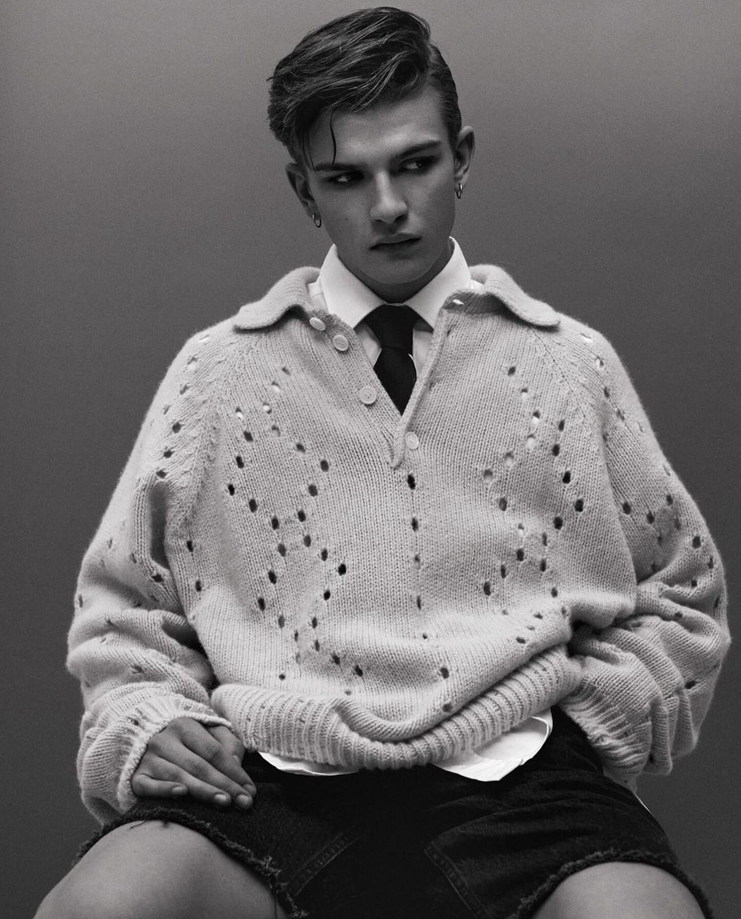 Portrait of a young adult male in an off-white open-knit sweater, white collared shirt, dark tie, and denim shorts, posing in a studio.