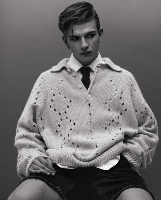 Portrait of a young adult male in an off-white open-knit sweater, white collared shirt, dark tie, and denim shorts, posing in a studio.