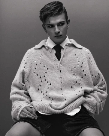 Portrait of a young adult male in an off-white open-knit sweater, white collared shirt, dark tie, and denim shorts, posing in a studio.