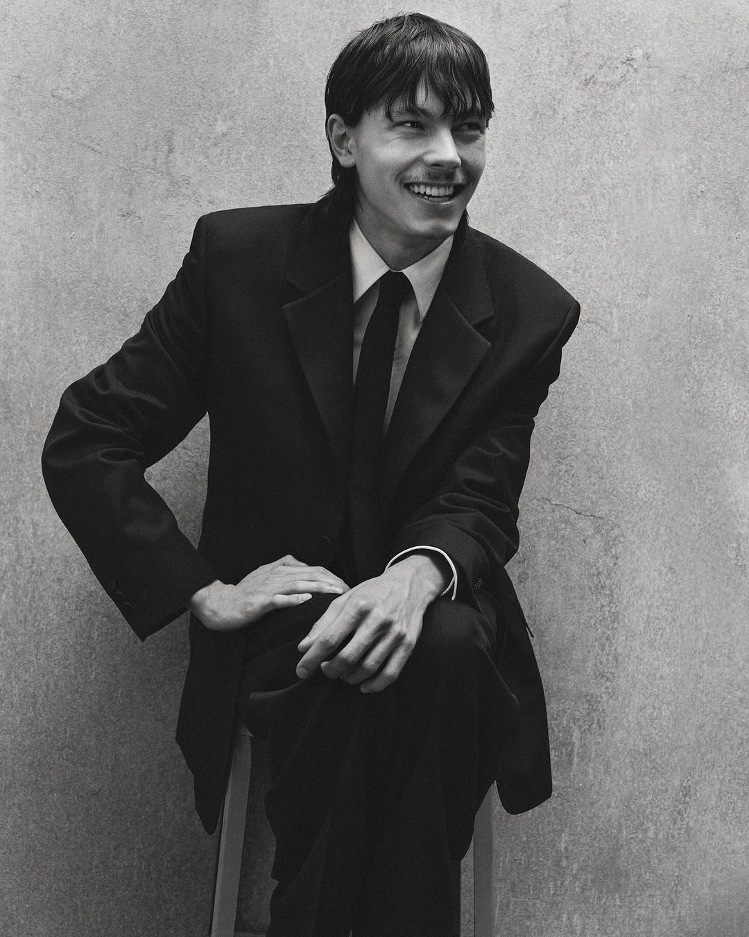 A smiling young man in a dark suit and tie seated against a textured concrete wall, captured in monochrome portraiture.