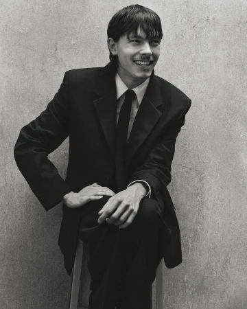 A smiling young man in a dark suit and tie seated against a textured concrete wall, captured in monochrome portraiture.