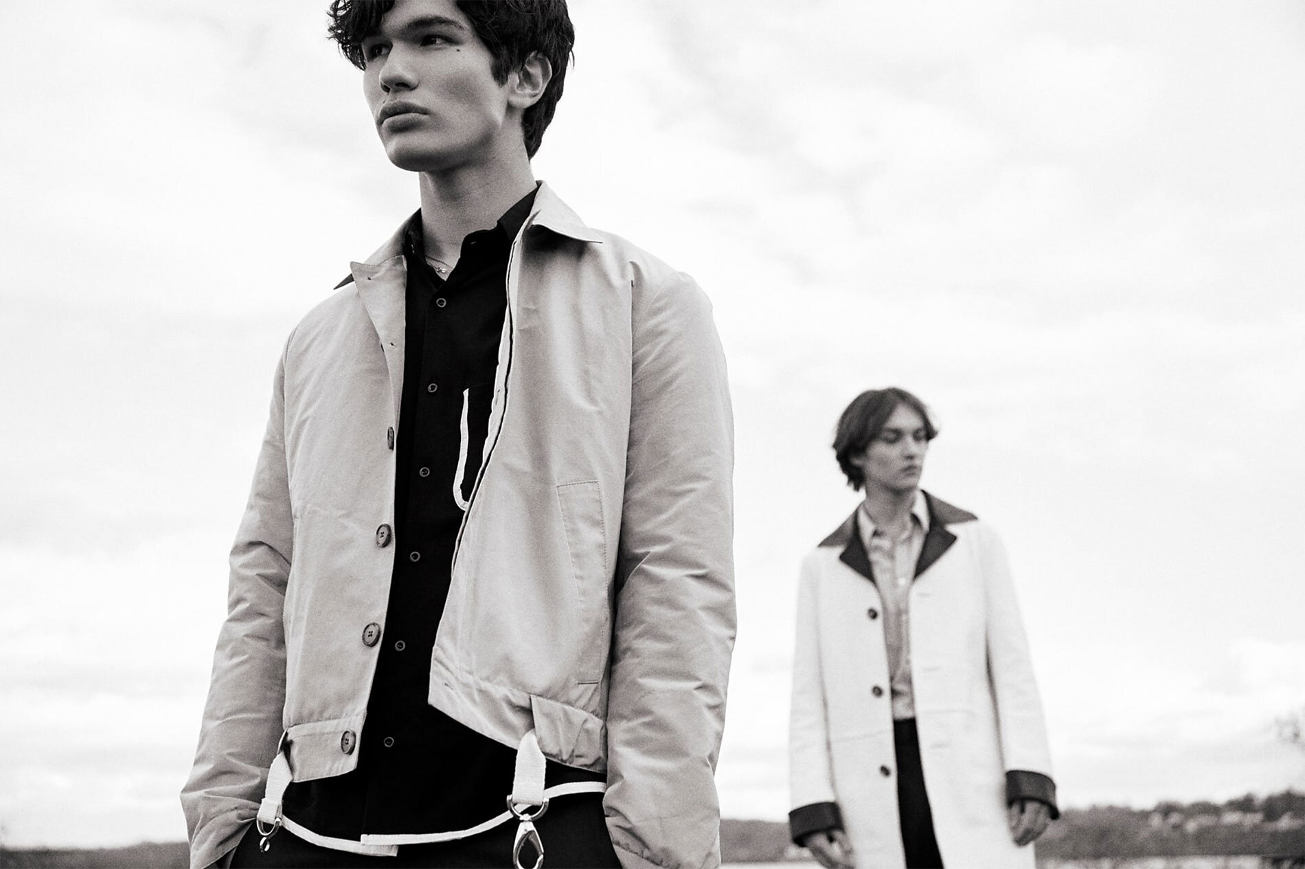 Black and white shot of two adult males in light jackets and dark shirts, standing outdoors against a cloudy sky. Fashion editorial.