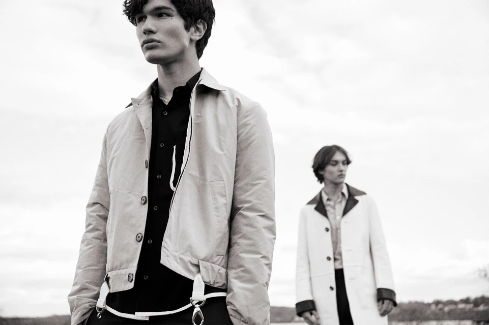 Black and white shot of two adult males in light jackets and dark shirts, standing outdoors against a cloudy sky. Fashion editorial.