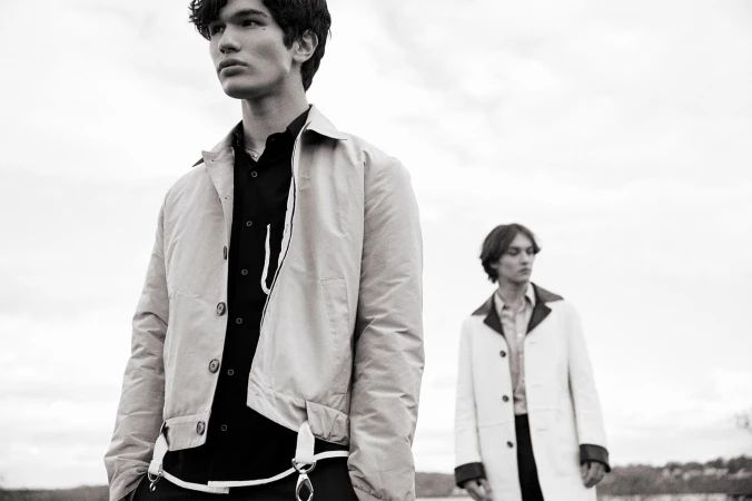 Black and white shot of two adult males in light jackets and dark shirts, standing outdoors against a cloudy sky. Fashion editorial.